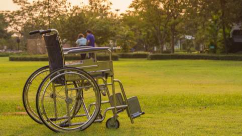 Foto: Im Vordergrund steht ein leerer Rollstuhl auf einer großen, grünen Wiese. Im Hintergrund, weit entfernt sieht man ein Paar 