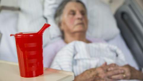 Im Vordergrund eine Schnabeltasse auf einem Beistelltisch. Im Hintergrund liegt eine ältere Frau in einem Pflegebett.