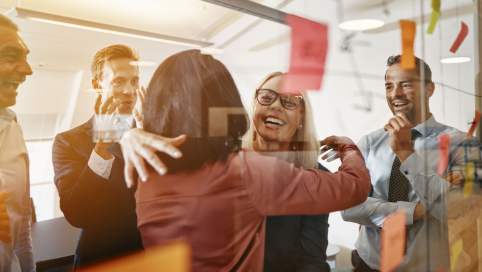 Foto: Hinter einer Glaswand sieht man wie eine Mitarbeiterin vor anderen Kollegen von einer Kollegin herzlich begrüßt und umarmt wird